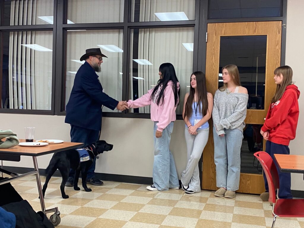 A man holds the leash of a black lab as he shakes hands of a teenager. Three more teenagers are lined up behind them, and the black lab is wearing a Future Leader Dog vest.