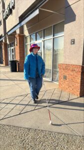 Wearing a blue coat, jeans and a great, red hat, a woman uses her long white cane to travel on a sidewalk.