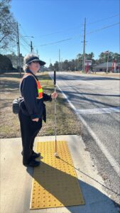 Dressed in black with a yellow safety vest, a woman stands on yellow truncated domes holding a long white cane while waiting for the light to change.