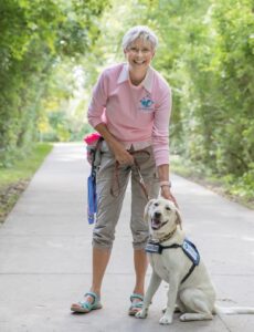 A woman with short white hair stands next to a yellow lab. They are in an outdoor trail on a sunny day, surrounded by greenery. The woman wears a treat pouch and holds the dog's leash.