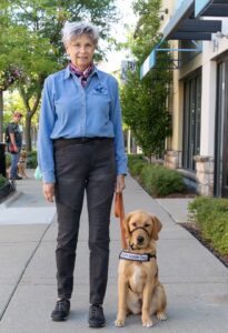 A woman with short grey hair and wearing a dress shirt and pants stands on a sidewalk on a sunny day. She holds the leash of a yellow lab puppy. The puppy is wearing a Future Leader Dog vest.