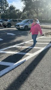 A person crosses the parking lot. They wear a pink shirt and jeans and is using their white cane.