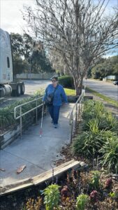 A person walks on a ramp. They are using their white cane and is wearing a blue shirt and jeans.