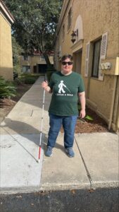 A person stands on the sidewalk outside their home. They are wearing jeans and a green Leader Dog shirt depicting the figure of a person using a cane. The caption reads “I drive a stick”.