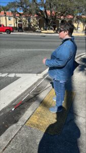 A person stands at the intersection, preparing to cross the street. They wears jeans and a blue shirt and is holding their white cane in her right hand.
