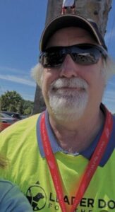 A man wearing a ballcap, sunglasses and a medal smiles toward the camera on a sunny day.