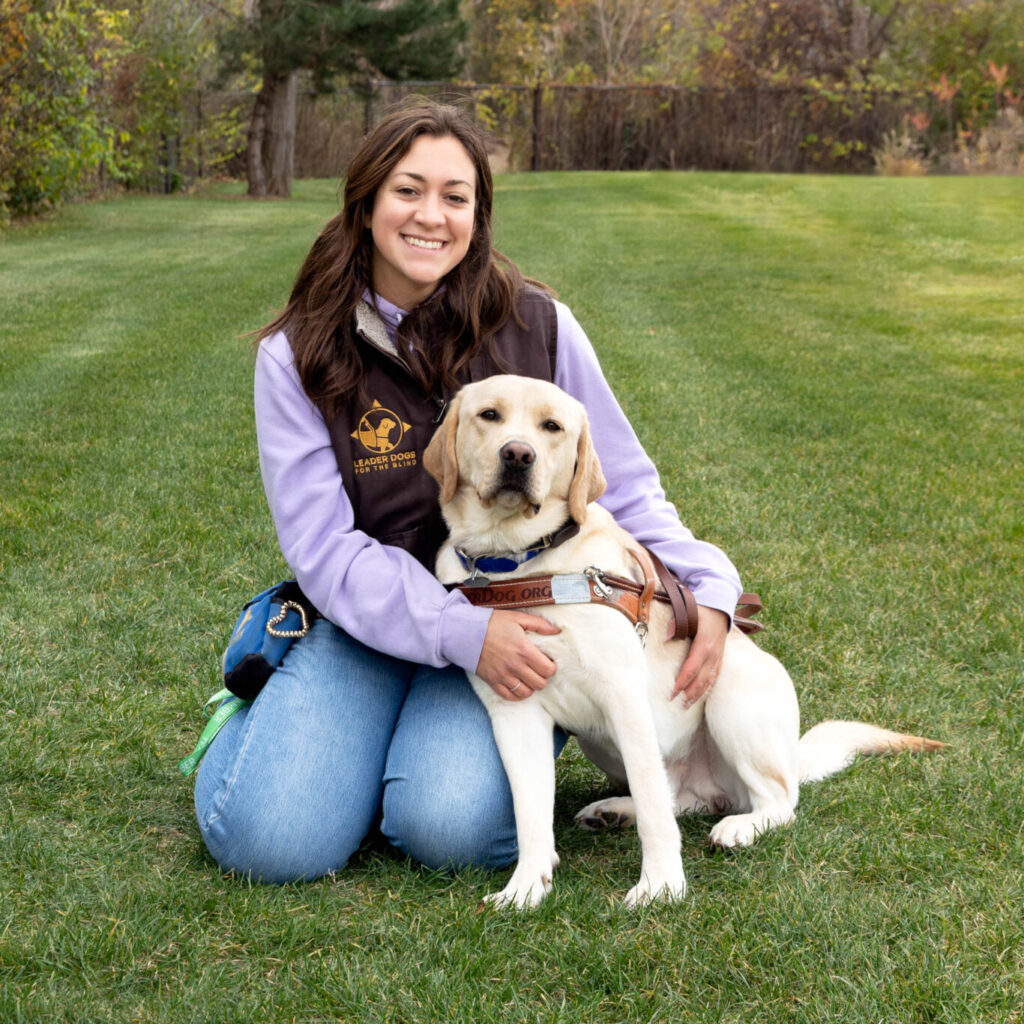 A person kneels on green grass in an open park, holding a yellow Labrador retriever wearing a working harness. The person is dressed in a long-sleeve shirt, a dark vest, and jeans, with trees showing autumn foliage in the background.