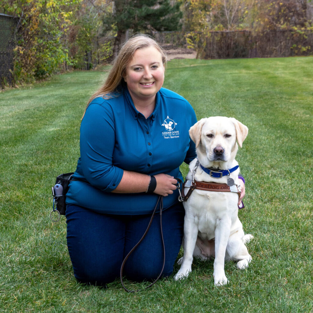 A person kneels on a grassy lawn beside a yellow Labrador retriever wearing a working harness and leash. The person is dressed in a teal long-sleeve top and dark pants, with trees showing autumn colors in the background of an open outdoor setting.