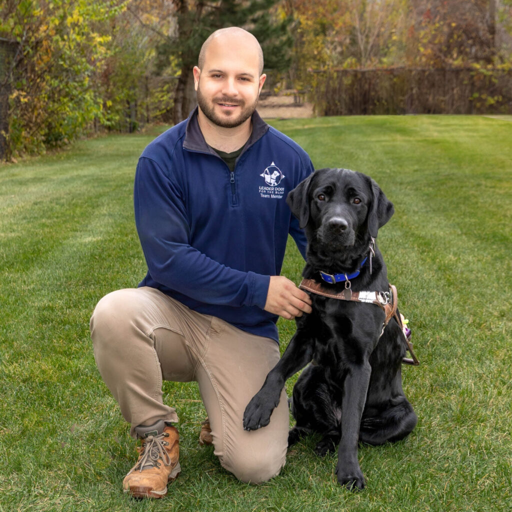 A person kneels on a neatly mowed grassy lawn beside a black Labrador retriever wearing a working harness and blue collar. The person is dressed in a navy long-sleeve top, khaki pants, and brown boots, with one hand resting near the dog’s harness. Trees with autumn foliage and an open green space fill the background.