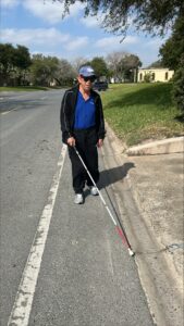Wearing a dark track suit and blue shirt, a man uses his cane to trail the curb on the left side of the street.