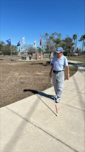 Wearing a light blue shirt, gray pants, and sunglasses, a man uses his white cane to travel on the sidewalk at Veterans Park. Flags from each branch of the U.S. Military are in the background against a beautiful, blue sky.