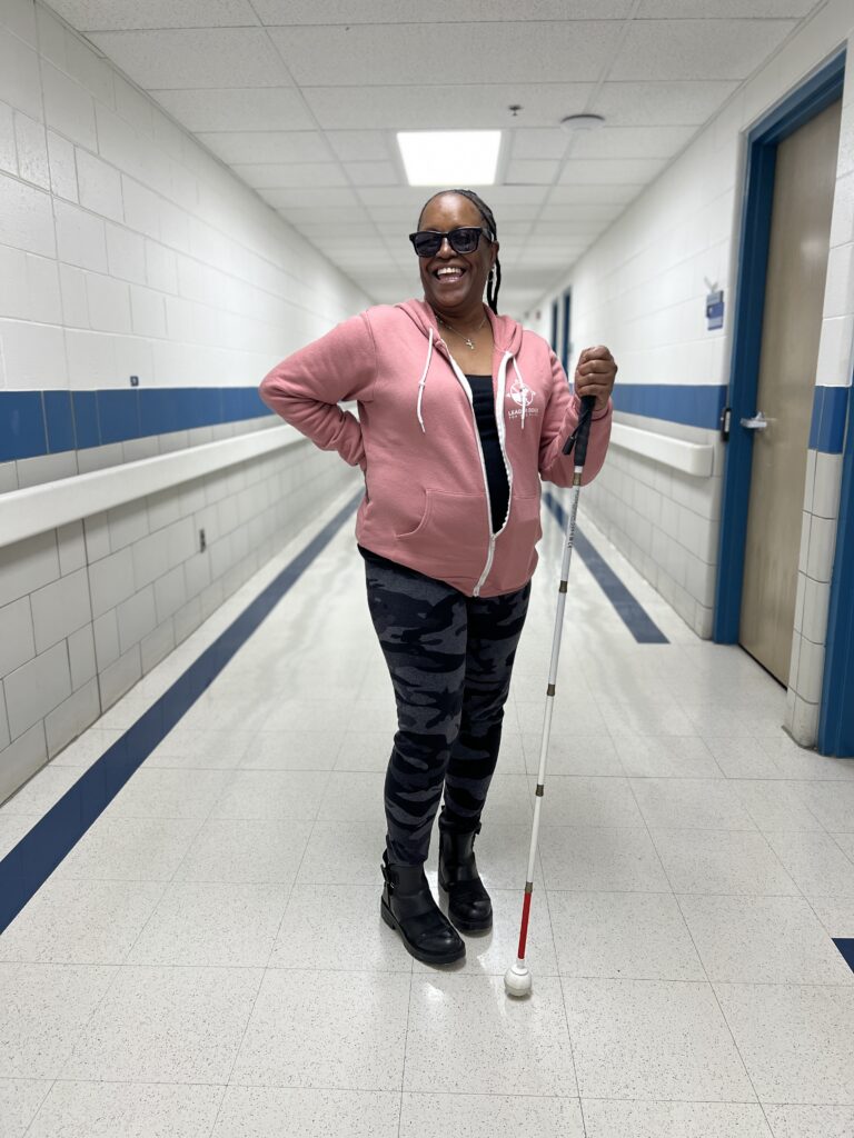 A person stands in a long, well-lit hallway with white and blue walls, holding a white mobility cane with a red tip. The person is wearing a pink zip-up hoodie, dark patterned pants, and black boots, with one hand resting on the hip and doors lining the corridor in the background.