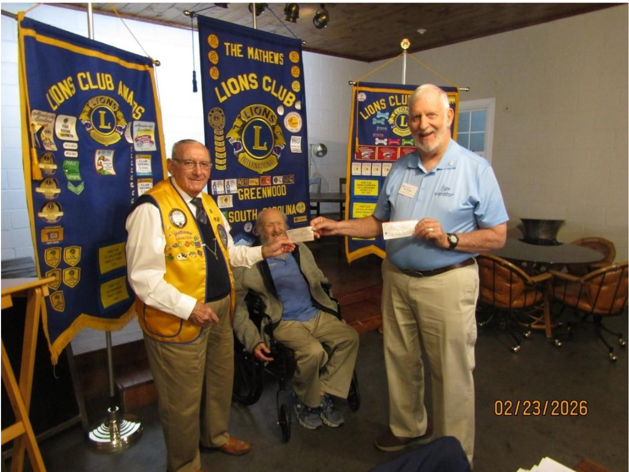 Three smiling men standing in front of Lions Club banners. One is sitting down in the middle, and two standing and holding a check between them.