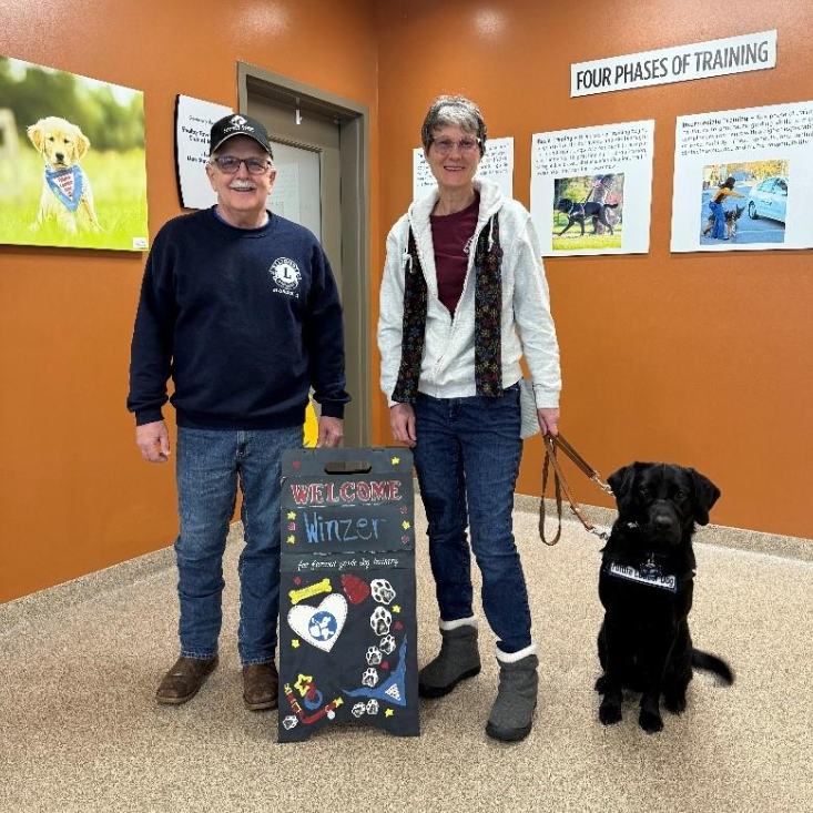 A couple stands smiling with their future Leader Dog, a black lab. They are posing with a sign that says "Welcome Winzer"