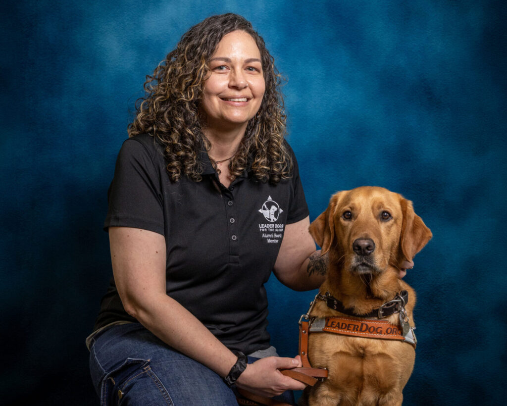 A woman with dark curly hair smiles toward the camera. Next to her is a yellow Lab Leader Dog in harness, also looking toward the camera.
