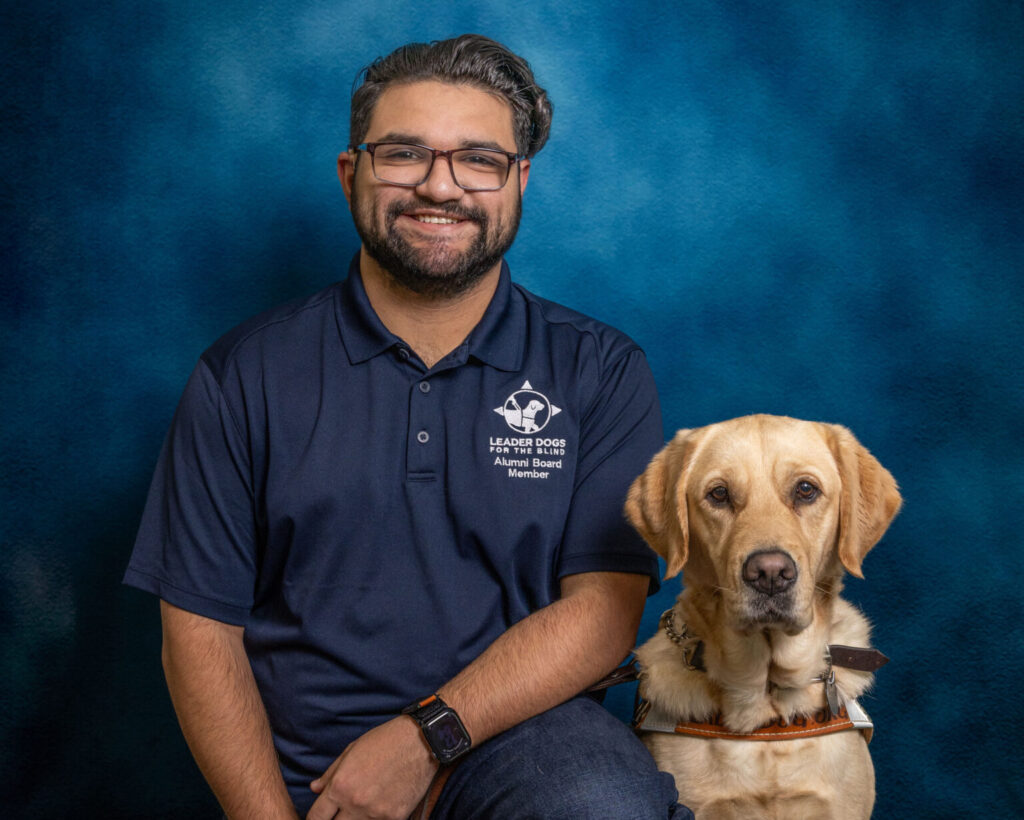 A man with dark hair and glasses smiles toward the camera. Next to him is a yellow Lab Leader Dog in harness, also looking toward the camera.