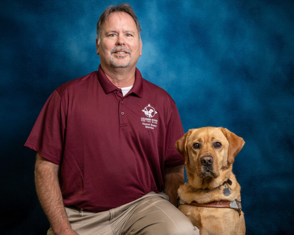A man with light colored hair and a goatee smiles toward the camera. Next to him is a yellow Lab Leader Dog in harness, also looking toward the camera.