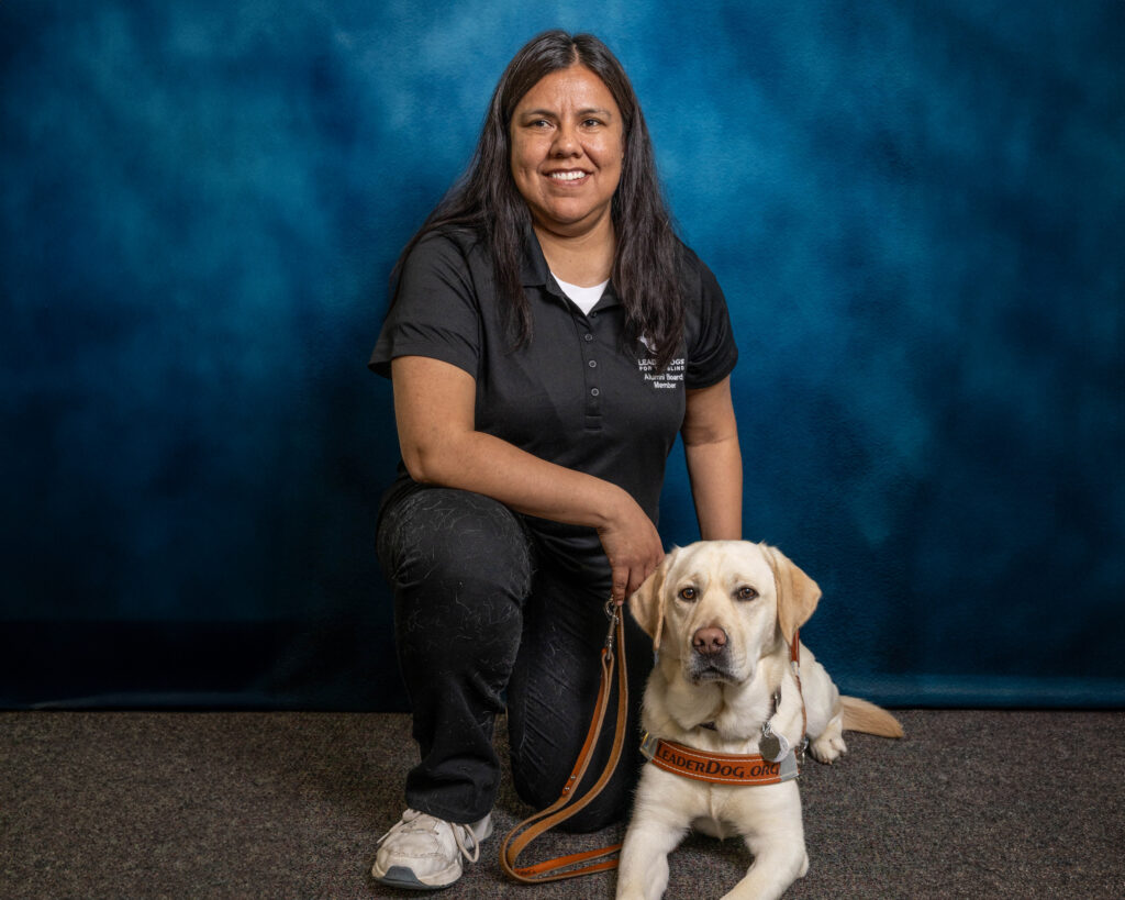 A woman with long black hair smiles toward the camera as she kneels holding the leash of the yellow Lab Leader Dog next to her. The dog also looks toward the camera.