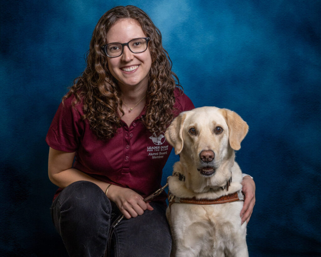 A woman with brown curly hair and glasses smiles toward the camera. Next to her is a yellow Lab Leader Dog in harness, also looking toward the camera.