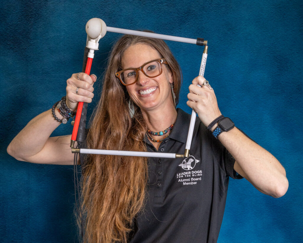A woman with long brown hair smiles toward the camera while holding a white cane folded into a square in front of her face like a frame.
