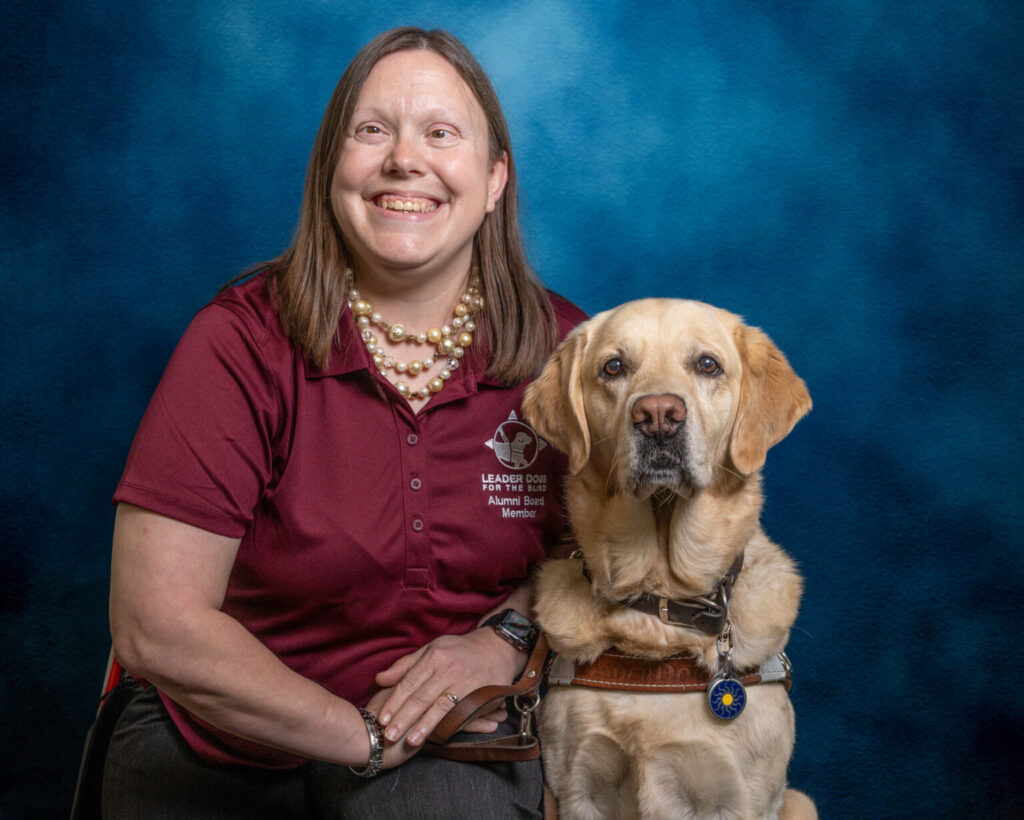 A woman with short, light brown hair smiles brightly toward the camera. Next to her is a yellow Lab Leader Dog, also looking toward the camera.
