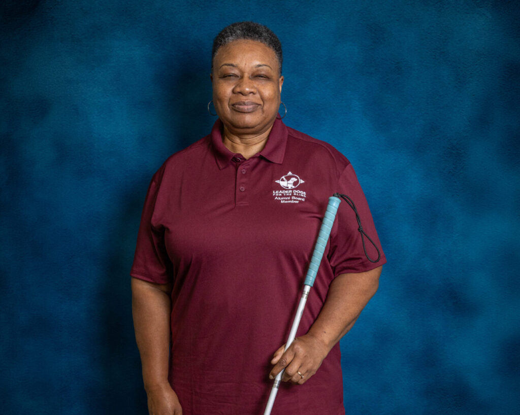 A woman smiles toward the camera. She has short curly hair and is wearing hoops. She holds a long white cane where the handle is a teal color.