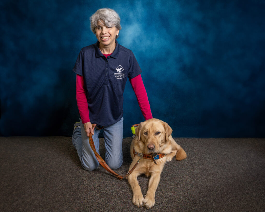 A woman with short grey hair kneels as she smiles toward the camera. Next to her is a yellow Lab Leader Dog in harness. She holds the guide dog's leash in one hand and pats him with the other.