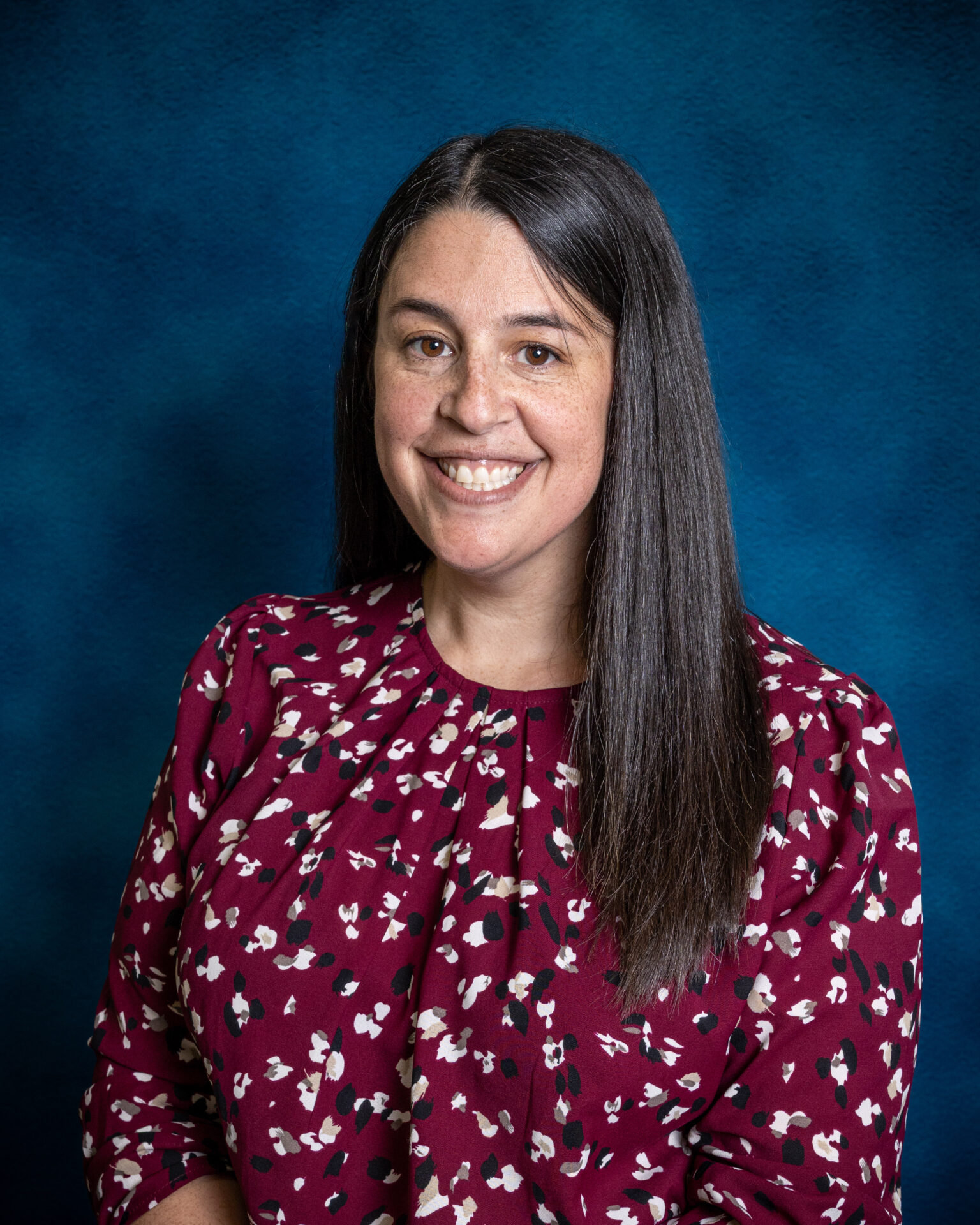 A smiling headshot of a woman with long dark hair in a patterned burgundy blouse against a dark blue backdrop