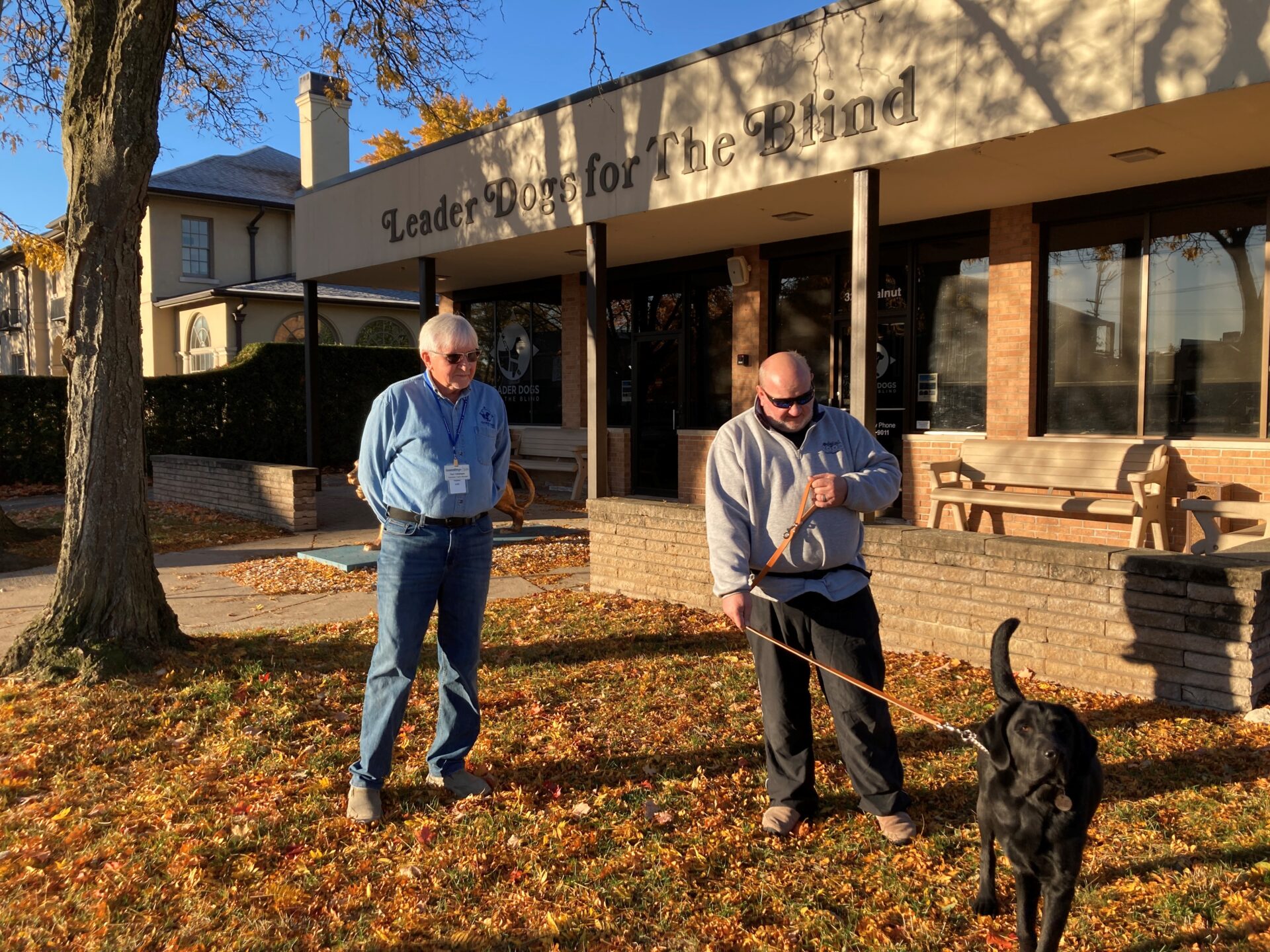 Two men stand on a leaf-covered lawn with a black Lab outside a building labeled ‘Leader Dogs for the Blind.