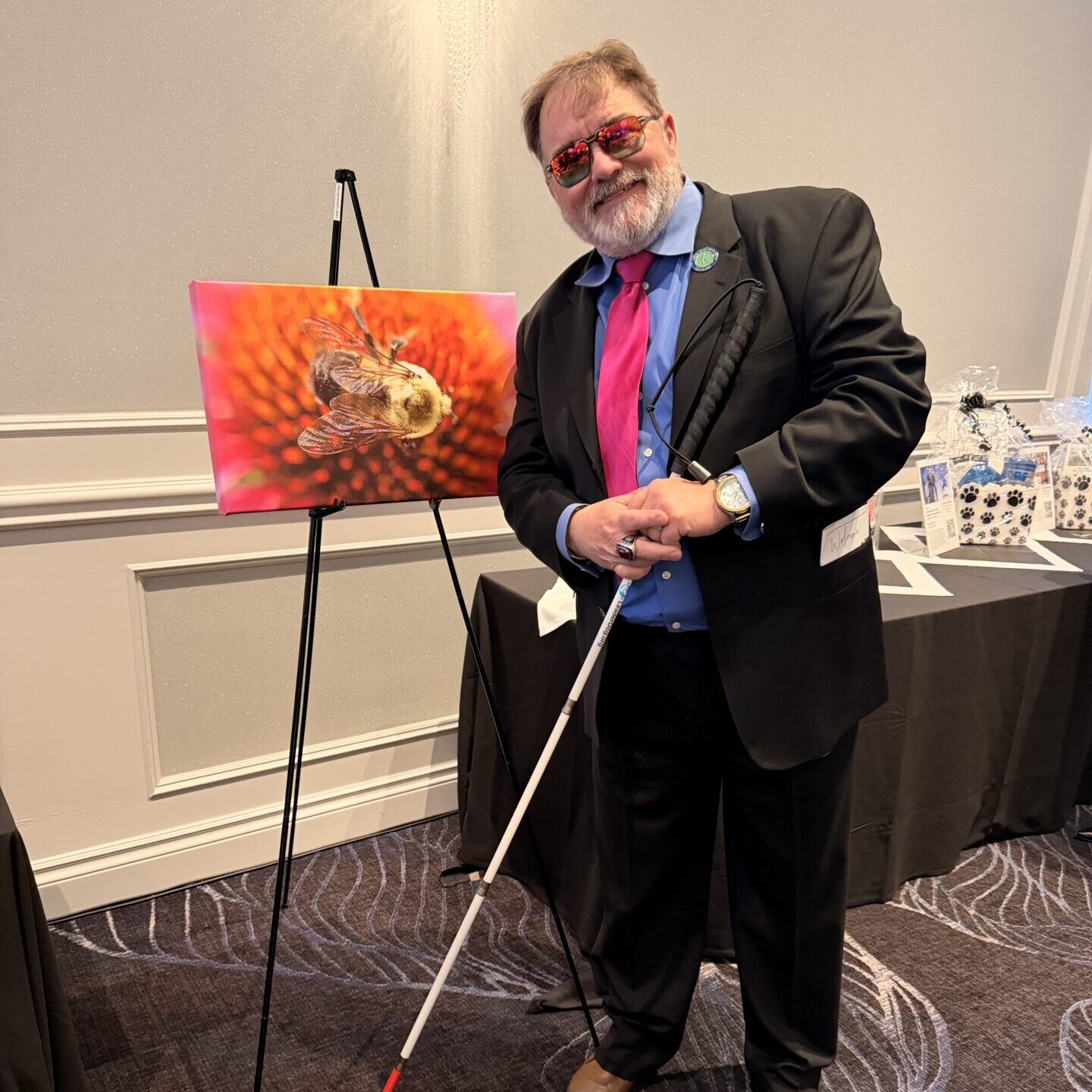 Man in a dark suit and bright tie smiles while holding a white cane, standing beside a framed colorful artwork on an easel at an indoor event.