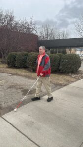 A man wearing a red jacket and khaki pants crosses a driveway using a white cane