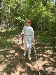 A woman hikes a wooded trail with a white cane in one hand and a trekking pole in the other.