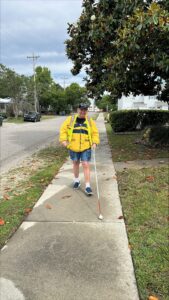 Wearing a hi-vis yellow jacket, a man travels on the sidewalk using his white cane.