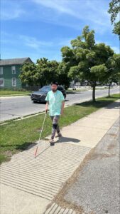 A man uses his white cane to navigate a sidewalk near a town street