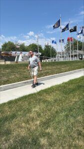 A man uses his white cane to navigate down a sidewalk, passing by flag poles flying assorted flags.