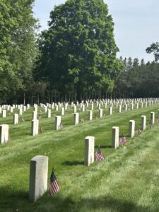 Rows of headstones, each with a small American flag, sit in neat rows in a grassy field