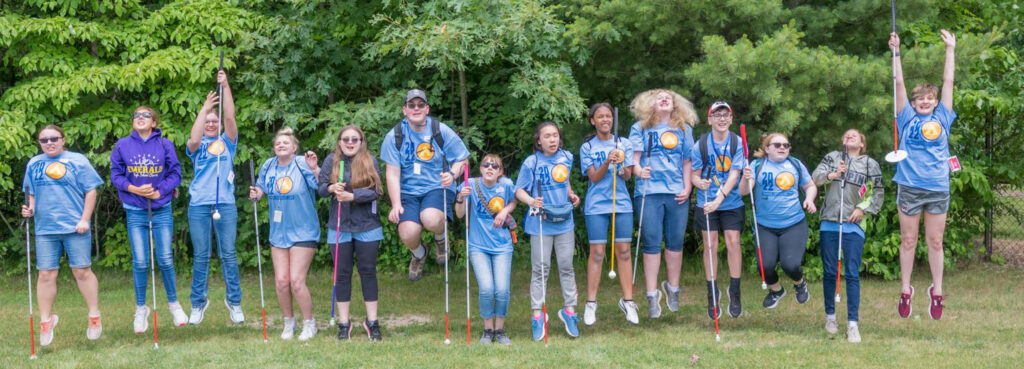 A group of teens with white canes stand outside the Leader Dog training facility. Several Leader Dog staff members pose with them as everyone smiles.