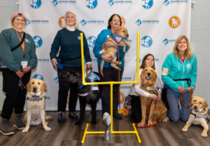 A group of women stand and smile in front of a Leader Dog branded step and repeat backdrop. Each woman has a dog in a Future Leader Dog vest with them. In front of the women is a yellow football goalpost.
