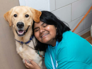 A teen girl with dark hair smiles as she leans into a yellow Labrador retriever.