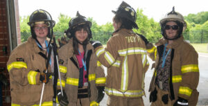 Four teens stand in a fire station smiling and wearing fire fighter gear with helmets. One of them faces away so you can see Troy on her back. Two are holding white mobility canes.