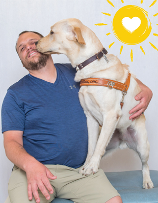 A man sits smiling as a yellow Labrador retriever in harness stands with its front paws on his leg and gently touches its nose to his cheek.