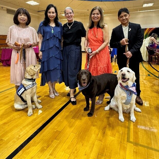 A group of Lions club members stand in a gymnasium with two retired Leader Dog Moms and one Future Leader Dog