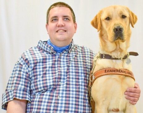 A man in a plaid button down sits proudly next to his guide dog, a yellow Lab in a leather harness that reads 