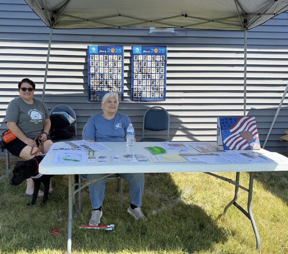 Two people sit at a table under an outdoor tent. The person on the left is holding the leash of a black Lab wearing a Future Leader Dog harness.