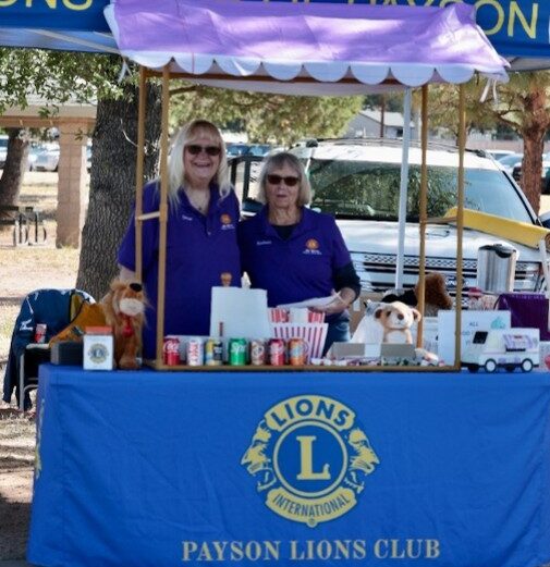 Two Payson Lions host a concession stand where they sell food and drinks for the event. The Payson Lions Club logo is printed on the tablecloth and tent covering.