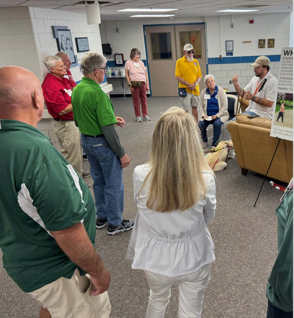 A group of people and volunteers stand in the lobby of the Polk Residence Center at Leader Dog’s campus. The group listens to a man speak while his Leader Dog rests at his feet.