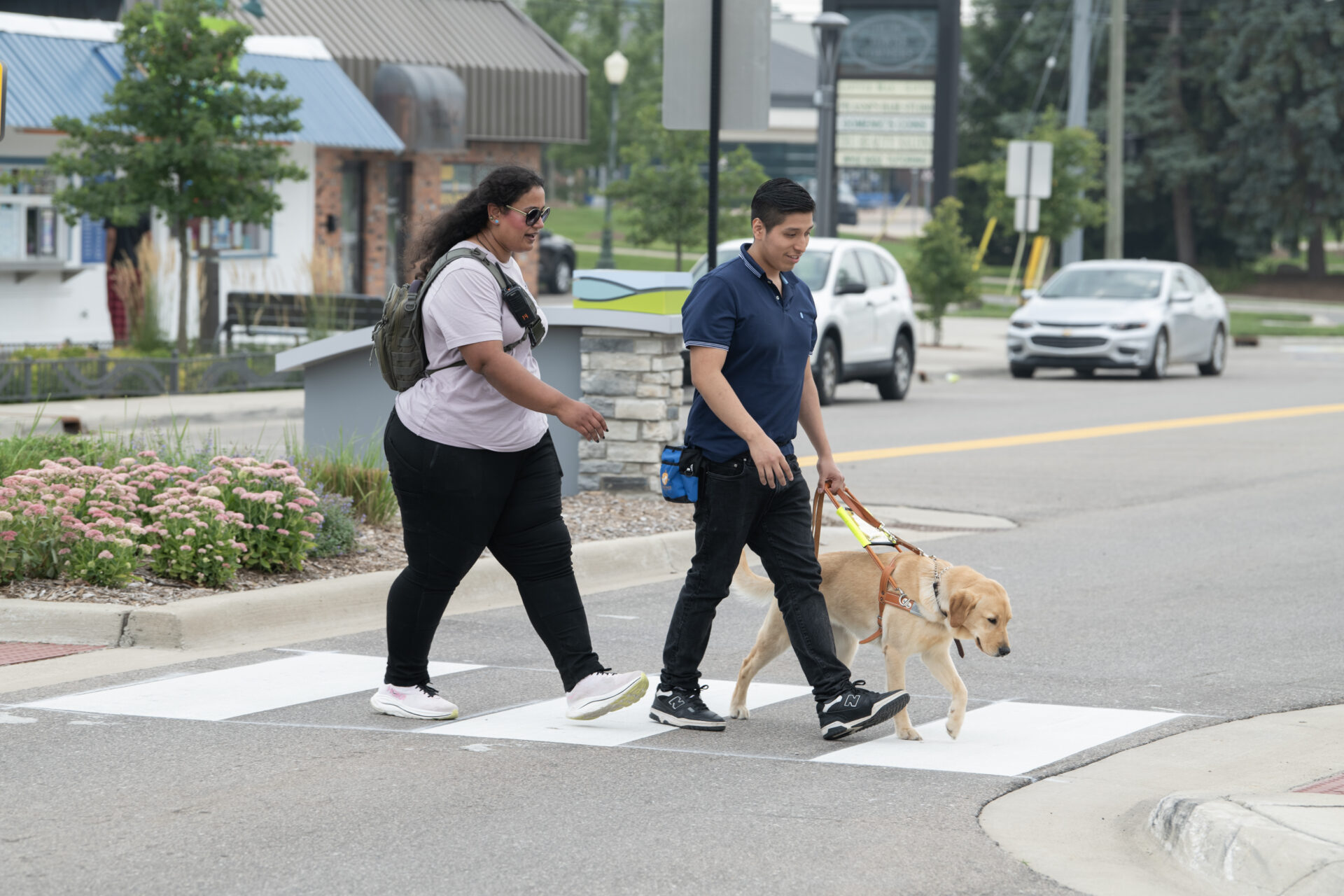 A man crosses the street with a yellow lab guide dog. A few steps away is a woman following them and appears to be giving instruction.