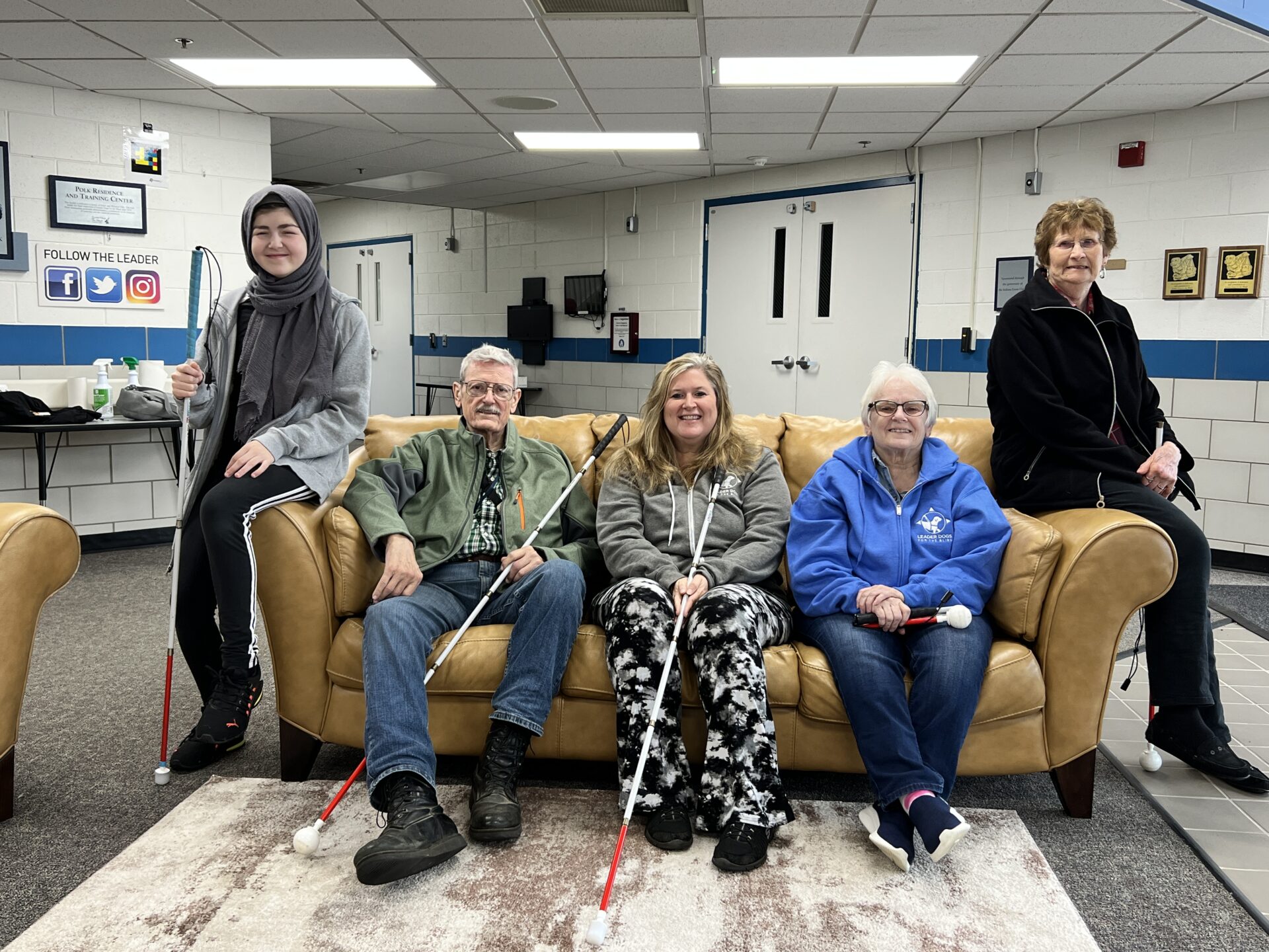 Five orientation and mobility program participants of varying ages pose with white canes around a couch in the Polk Residence lobby.