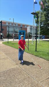 A woman uses her cane while walking down the sidewalk. She wears red top and denim skirt. In the background is an American flag and a bright blue sky.
