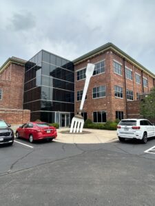 Large metal fork sculpture standing in front of a three-story brick office building with a glass entryway, surrounded by parked vehicles and landscaping under an overcast sky.